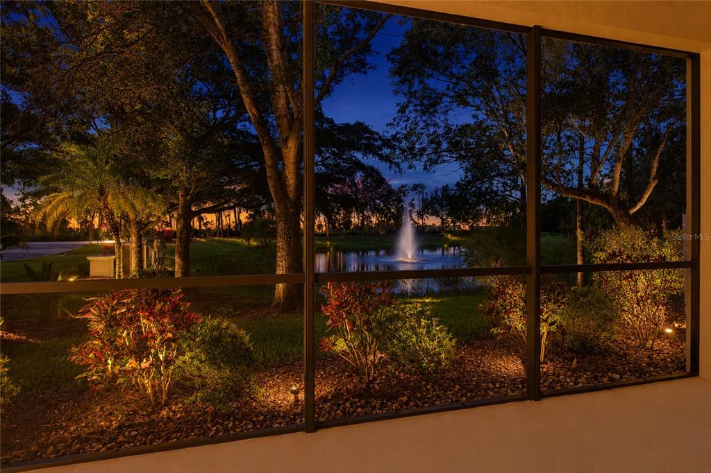 A view through a screened lanai at dusk showing a pond with a fountain and landscaping — 3781 COSTA MAYA WAY UNIT 101, ESTERO, FL