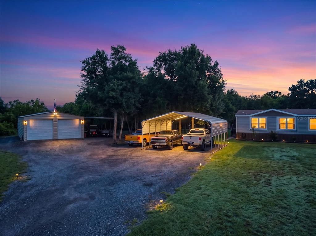An exterior view of a house with a detached garage and a metal carport at dusk — 353 URANUS TRAIL, OSTEEN, FL