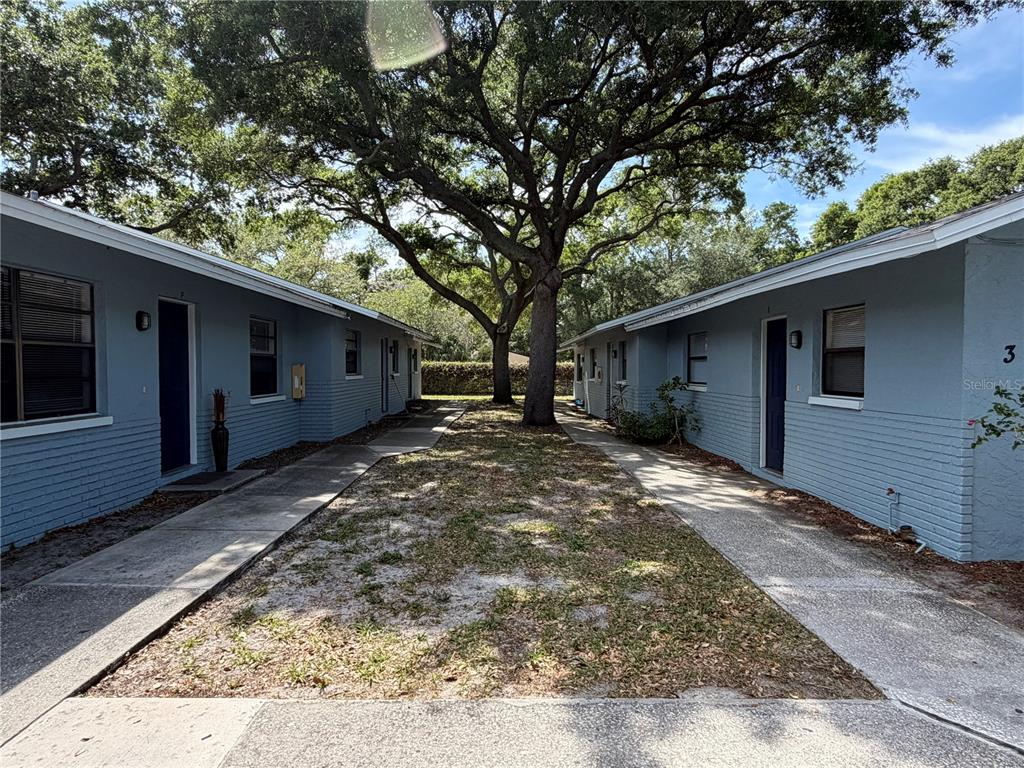 Two blue single-story apartment buildings face each other across a grassy courtyard with large trees — 950 BELTREES STREET, DUNEDIN, FL