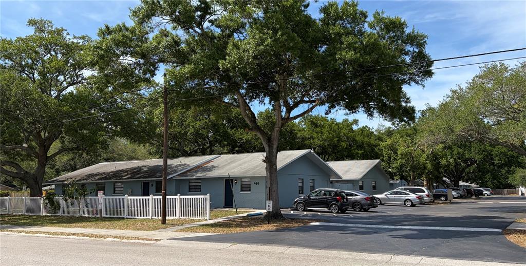 A single-story blue apartment building with a white fence and parking lot under large trees — 950 BELTREES STREET, DUNEDIN, FL