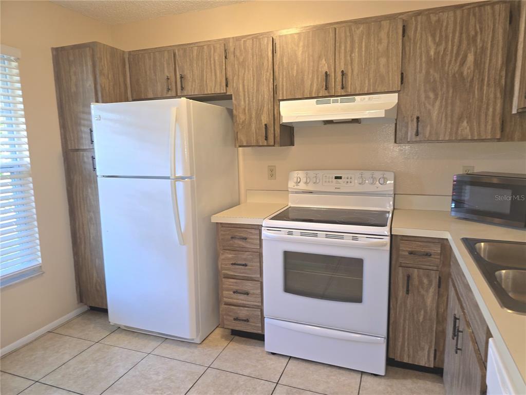 A kitchen features wood-grain cabinets, white appliances, a tiled floor, and a window with blinds — 2912 WAINWRIGHT COURT, NEW PORT RICHEY, FL