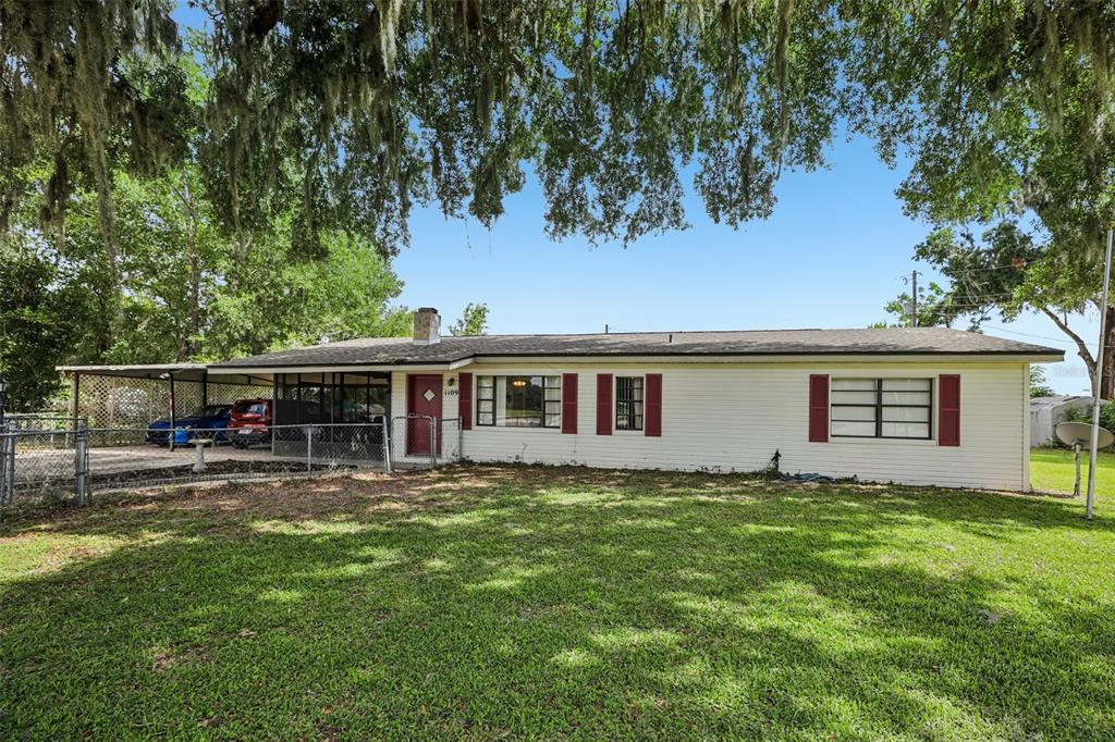 A single-story house with white siding, red shutters, and a covered carport under large trees — 1109 W C 48, BUSHNELL, FL