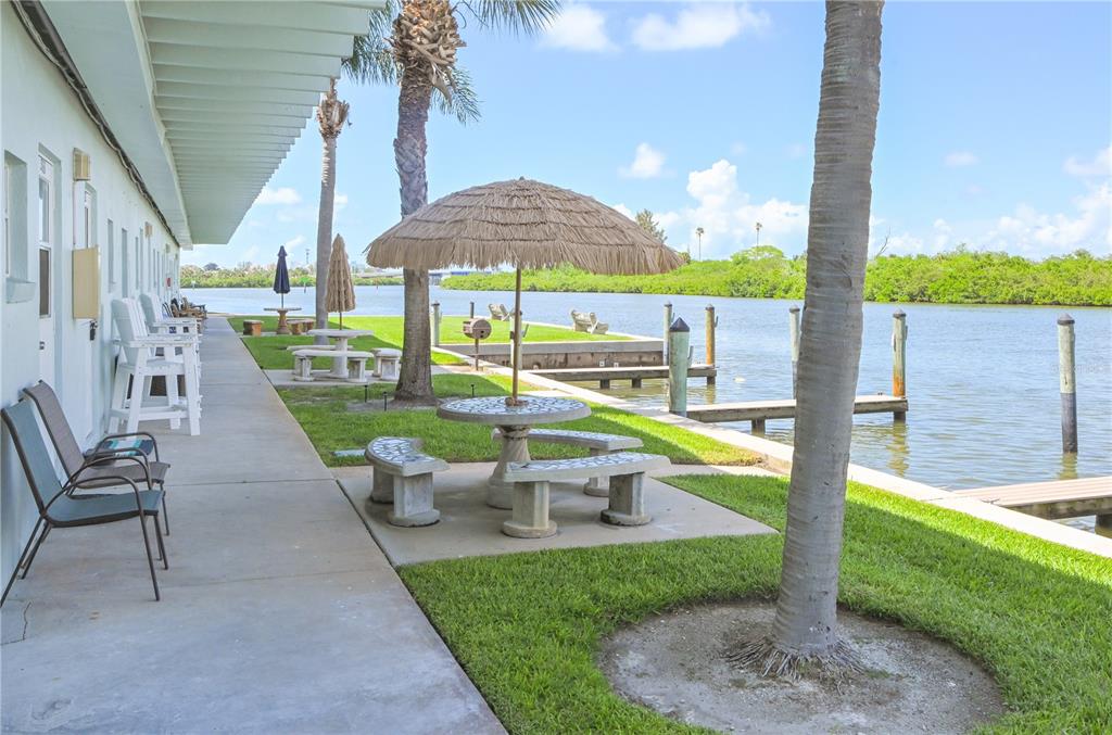A concrete patio with outdoor seating and a thatched umbrella overlooks a canal and dock — 18399 GULF BOULEVARD UNIT 367, INDIAN SHORES, FL