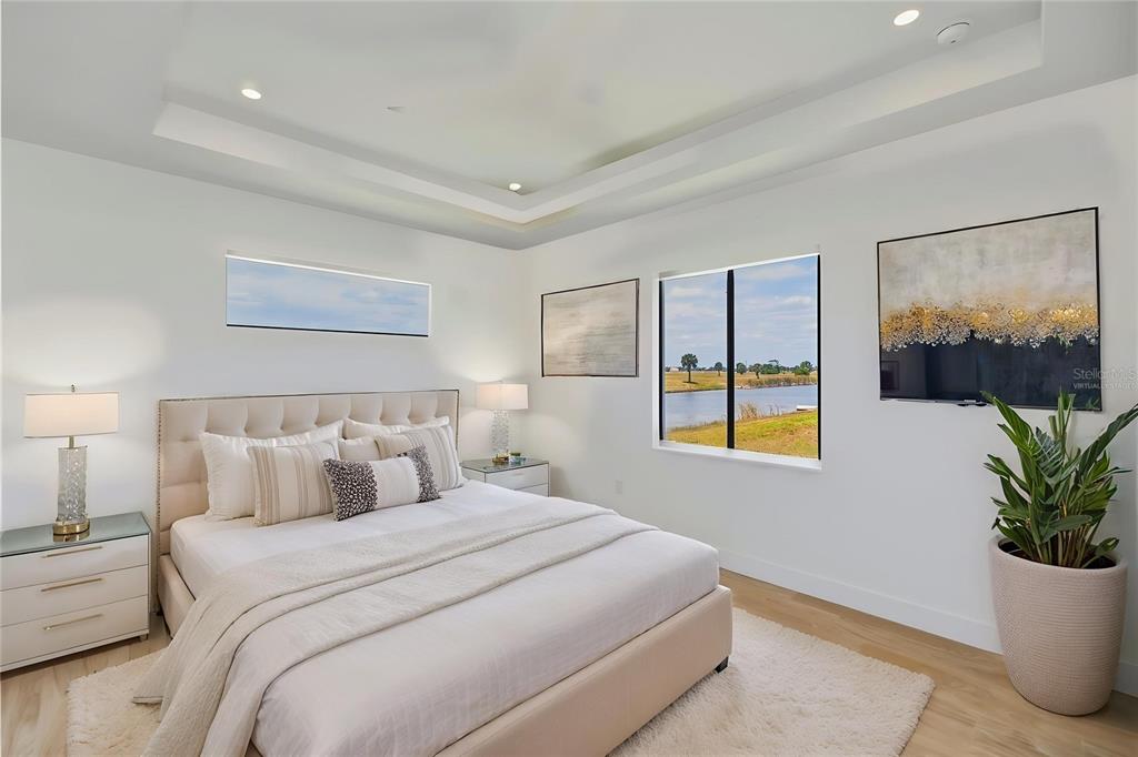 A modern bedroom features a tufted bed, tray ceiling, and a window overlooking a lake — 7 SUNFLOWER DRIVE, PLACIDA, FL