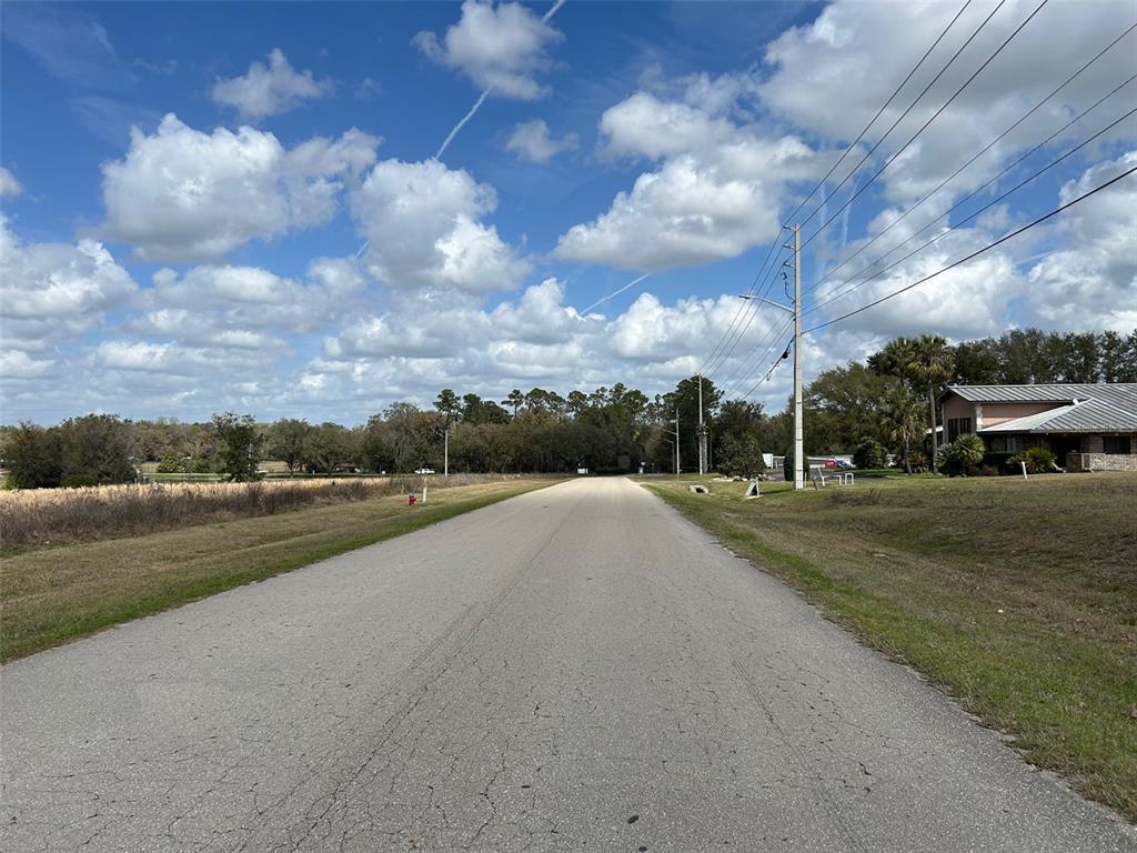 A paved road stretches toward a tree line under a bright, cloudy blue sky — NW 104TH TERRACE, ALACHUA, FL
