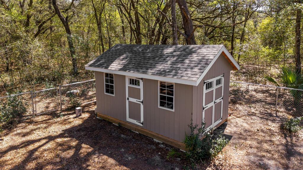 An elevated, tan-colored storage shed with a shingled roof sits in a wooded, fenced backyard — 23935 NE 118TH AVENUE, FORT MC COY, FL