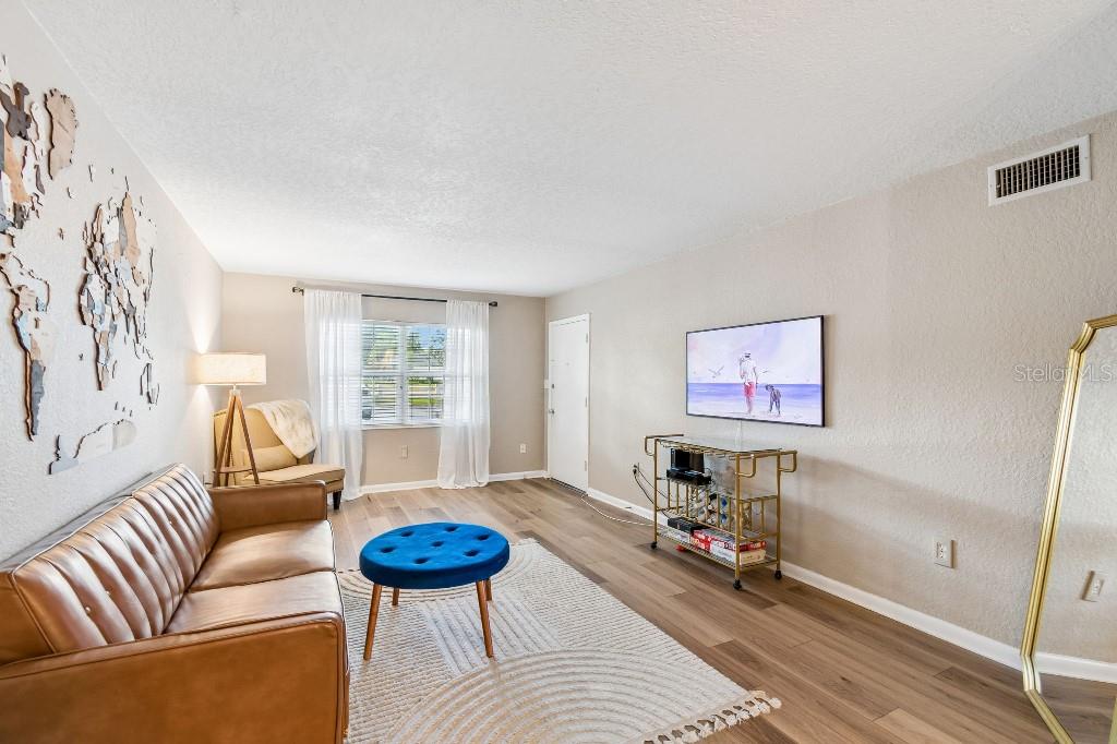 A living room features a brown leather sofa, blue ottoman, wall-mounted television, and wood flooring — 2500 LEE ROAD UNIT 104, WINTER PARK, FL