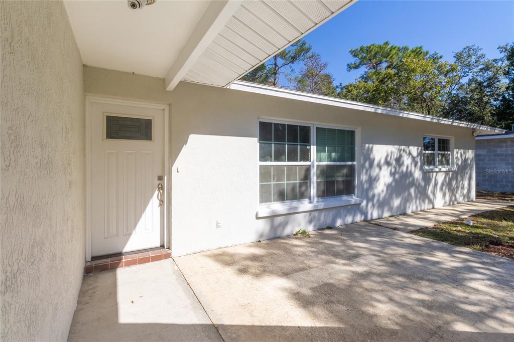 A white exterior wall features a front door, a large window, and a concrete patio — 6447 BRANDY DRIVE, SPRING HILL, FL
