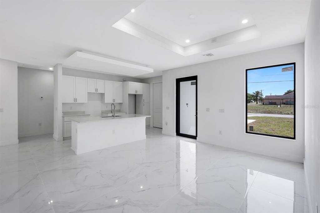 A modern, minimalist kitchen and living area with white cabinetry, marble-style flooring, and a tray ceiling — 100 PARISH DRIVE, LEHIGH ACRES, FL