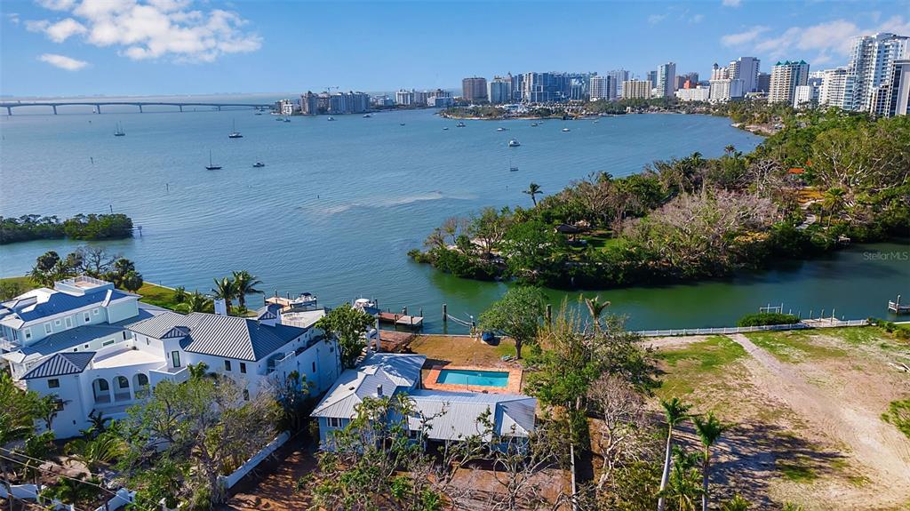 An aerial view shows a waterfront property with a pool, lush trees, and a city skyline — 1479 BAY POINT DRIVE, SARASOTA, FL