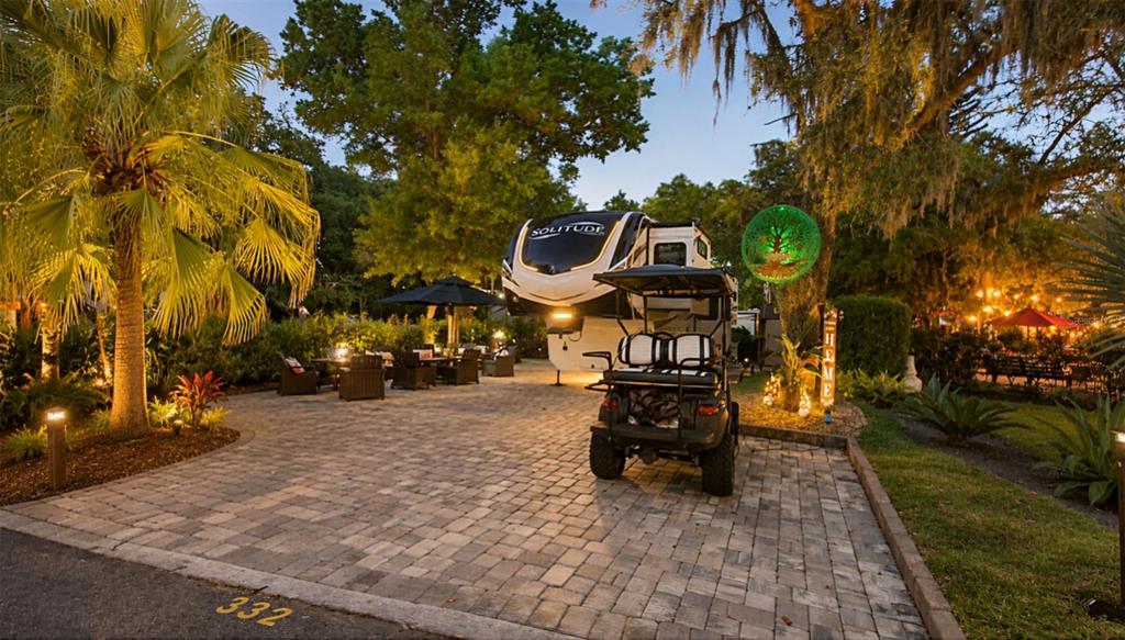 A landscaped yard features a hedge, several umbrellas, and mature trees under a bright sky — 332 BOBCAT LANE, LAKE WALES, FL