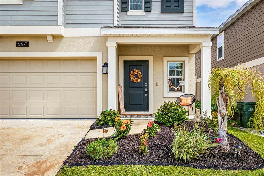 The exterior of a two-story house features a tan garage, dark door, and landscaped garden — 5571 BRAKEN FERN WAY, LEESBURG, FL