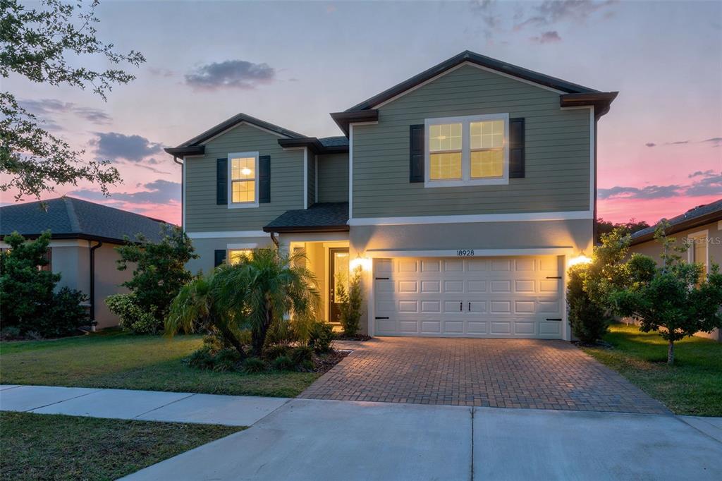 A two-story house with green siding, a brick driveway, and a two-car garage at dusk — 18928 WILLOWMORE CEDAR DRIVE, LUTZ, FL