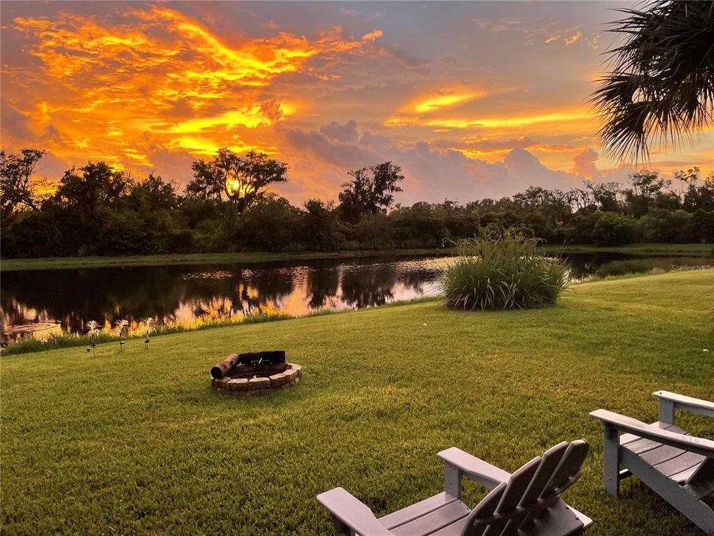 A grassy backyard features a stone fire pit, two chairs, and a pond at sunset — 6145 ANISE DRIVE, SARASOTA, FL
