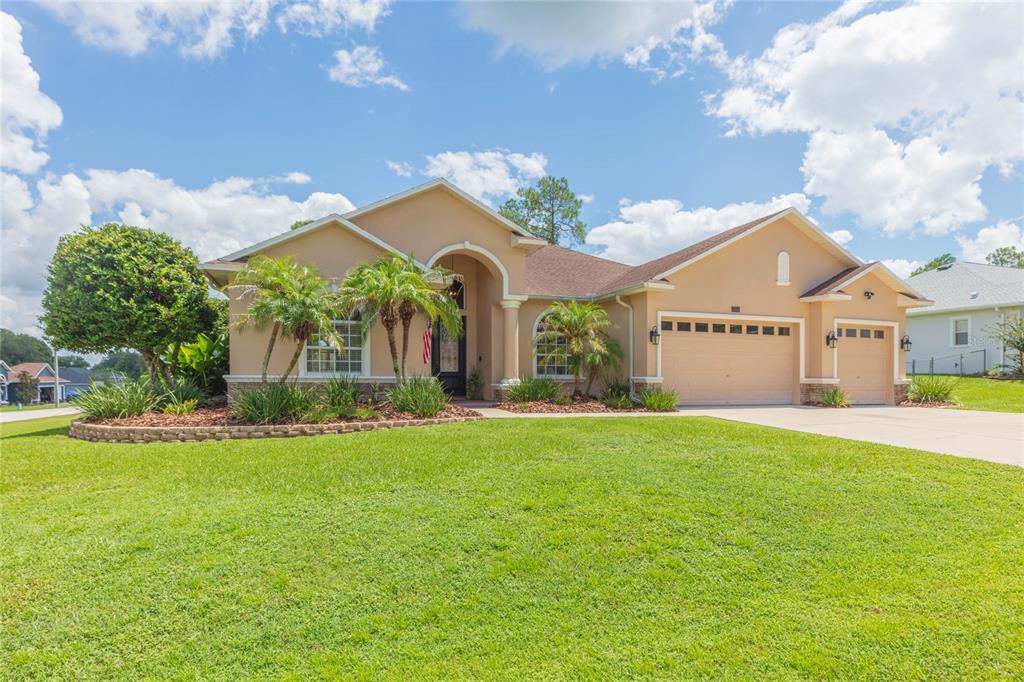 A single-story house with a tan stucco exterior, a three-car garage, and a landscaped lawn — 11544 PINE HOLLOW WAY, DADE CITY, FL