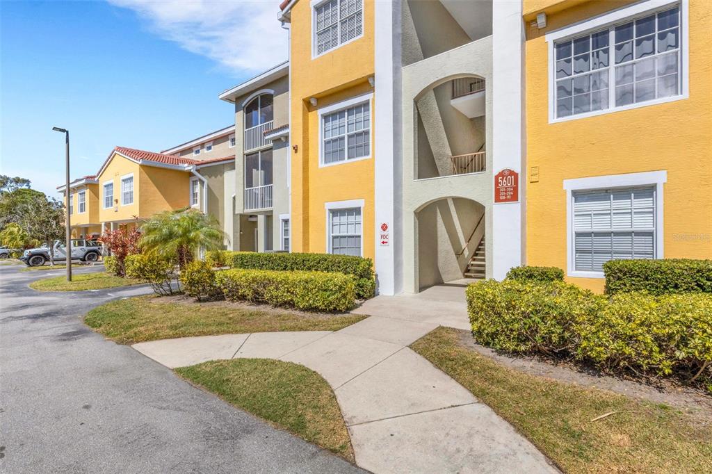 A three-story yellow apartment building with a concrete walkway and landscaped bushes in front — 5601 BENTGRASS DRIVE UNIT 10-202, SARASOTA, FL