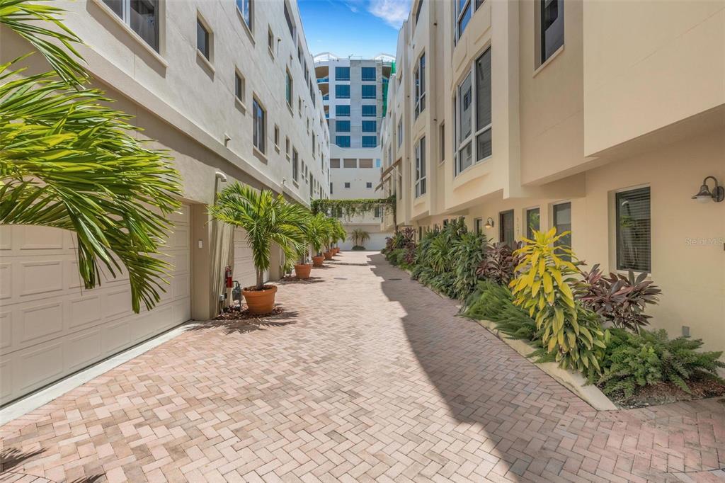 A paved alleyway between two multi-story residential buildings with potted palm trees and landscaping — 235 COCOANUT AVENUE UNIT 108B, SARASOTA, FL