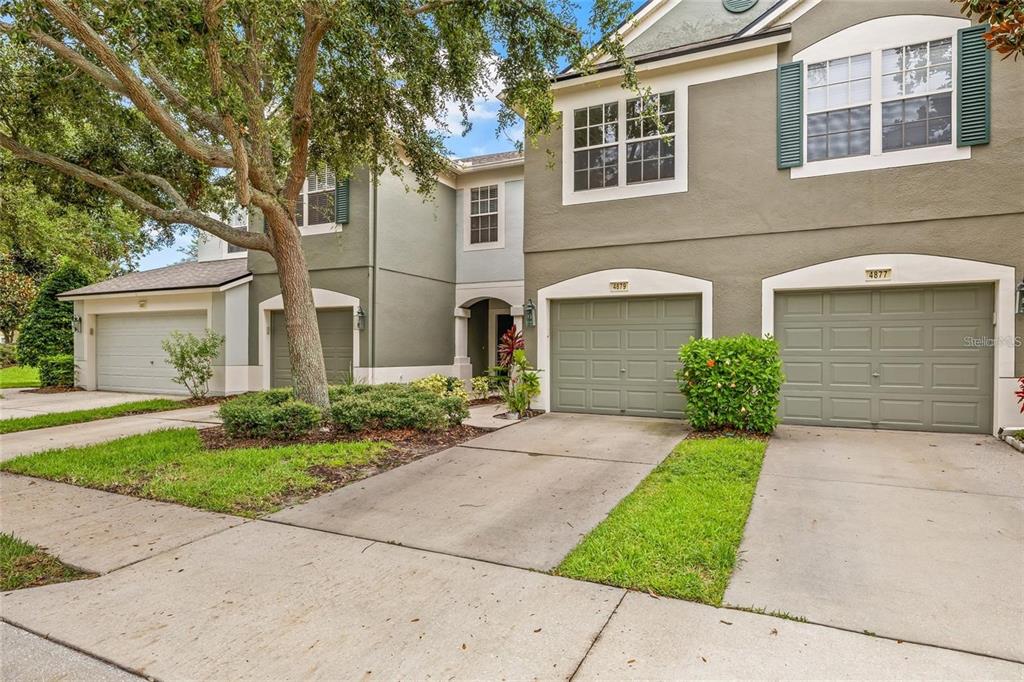 A two-story townhouse exterior features two garage doors, green shutters, and a large shade tree — 4879 POND RIDGE DRIVE, RIVERVIEW, FL