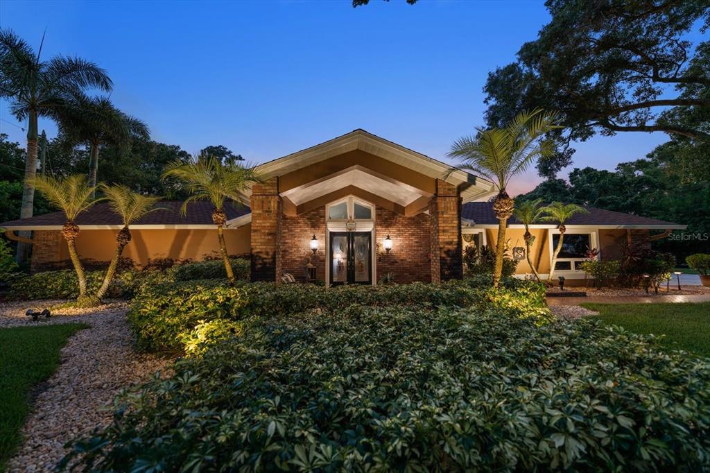 A brick-front ranch house with a gabled entryway, palm trees, and lush landscaping at dusk — 4141 WINDING WILLOW DRIVE, PALM HARBOR, FL