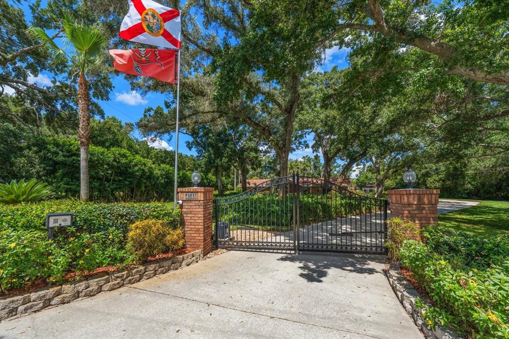 A gated driveway entrance features brick pillars, a metal gate, and two flags flying above — 4141 WINDING WILLOW DRIVE, PALM HARBOR, FL