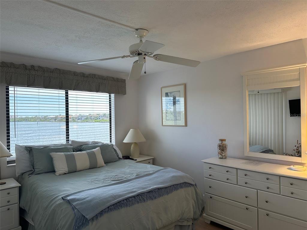 A bedroom features a bed, white dresser with mirror, ceiling fan, and a waterfront window view — 4835 BONITA BEACH ROAD UNIT 203, BONITA SPRINGS, FL