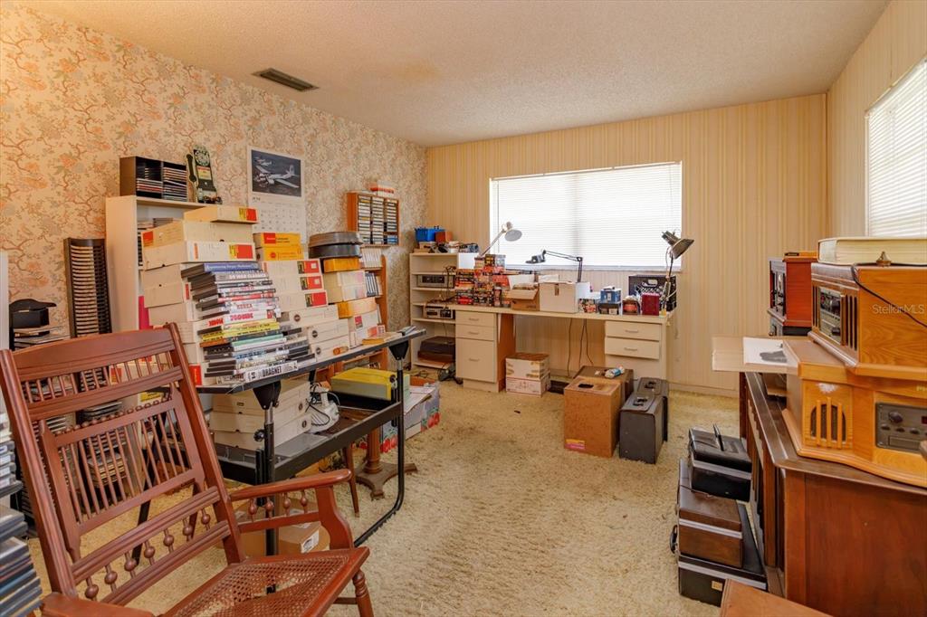 A cluttered home office features patterned wallpaper, beige carpet, and multiple desks with storage boxes — 1016 CATHY DRIVE, ALTAMONTE SPRINGS, FL