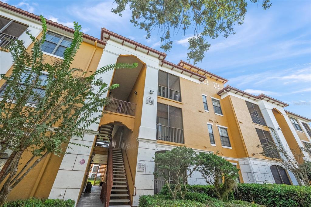 A three-story apartment building exterior features yellow stucco walls, screened balconies, and an outdoor staircase — 8341 38TH STREET CIRCLE E UNIT 303, SARASOTA, FL