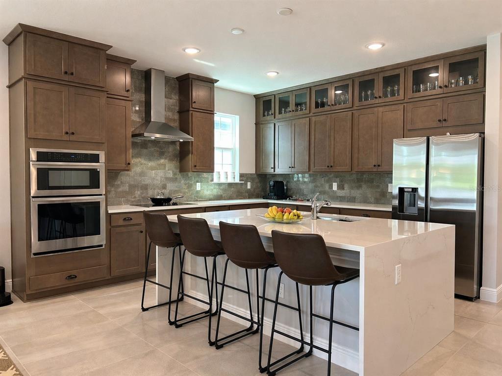 A modern kitchen features brown wood cabinetry, stainless steel appliances, and a large quartz island — 4891 SHADY PINES DRIVE, ST CLOUD, FL