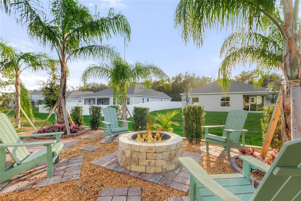 A backyard fire pit area features stone pavers, gravel, and green Adirondack chairs under palm trees — 2214 ELEGANT MANOR CIRCLE, EDGEWATER, FL