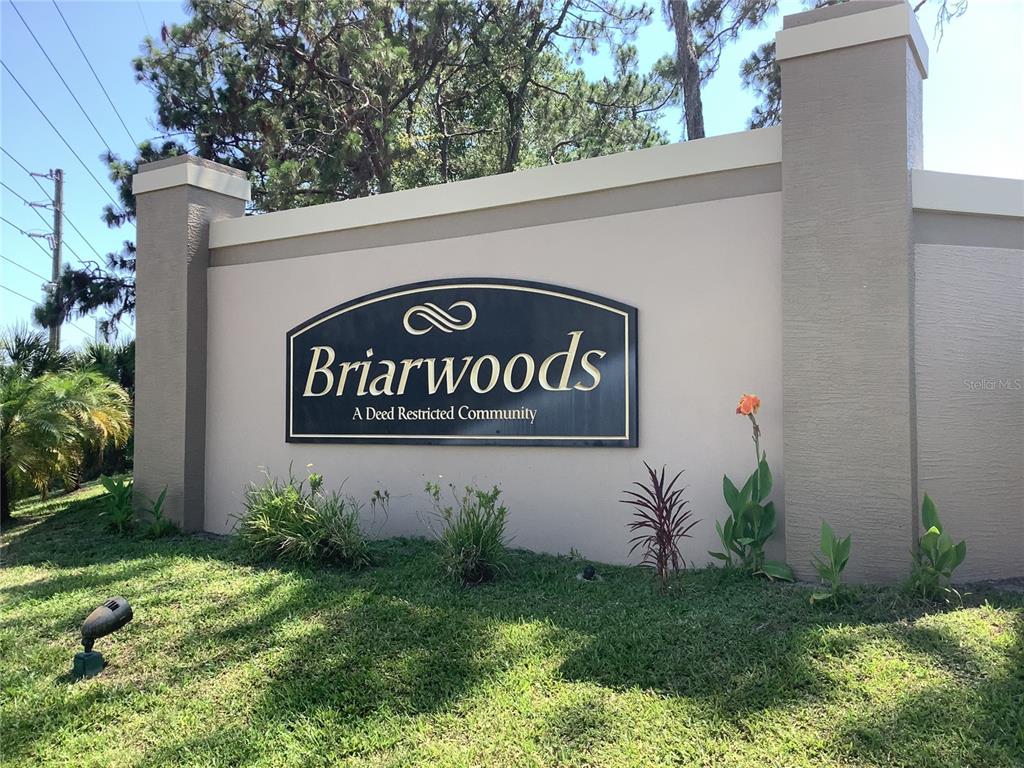 A beige stucco entrance sign for a community named Briarwoods stands in a grassy area — 18657 WHITE PINE CIRCLE, HUDSON, FL