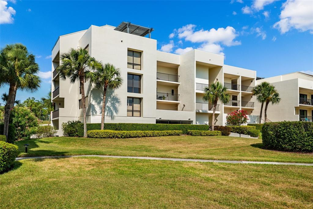 A multi-story white apartment building with balconies stands behind a grassy lawn and palm trees — 50 COE ROAD UNIT 331, BELLEAIR, FL