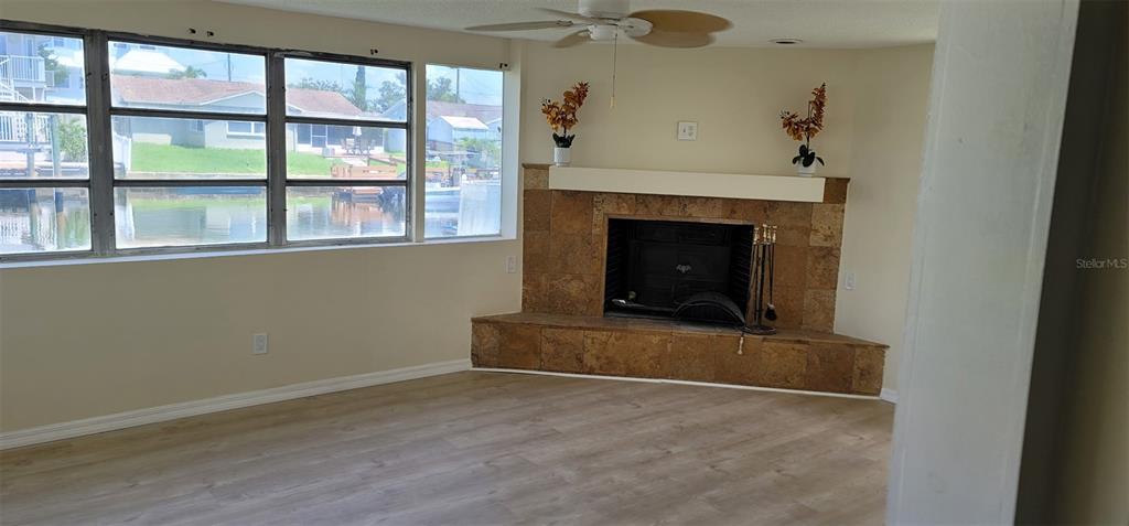 A living room features light wood-look flooring, a corner stone fireplace, and large windows overlooking water — 13240 STARFISH DRIVE, HUDSON, FL
