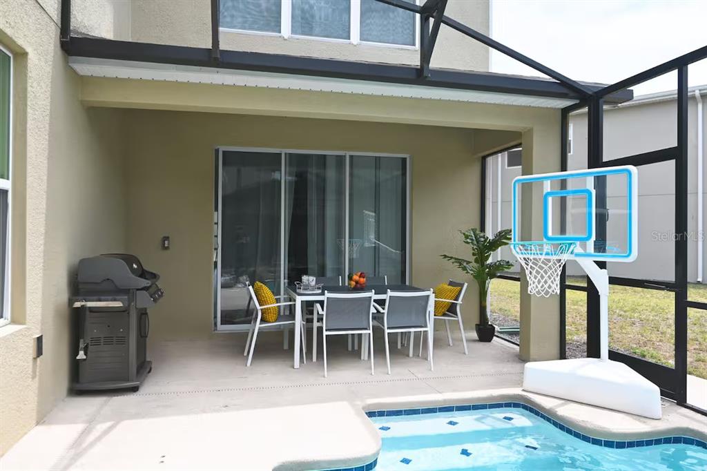 A covered patio features a dining set, a grill, and a basketball hoop by a pool — 2924 PROTAGONIST STREET, KISSIMMEE, FL