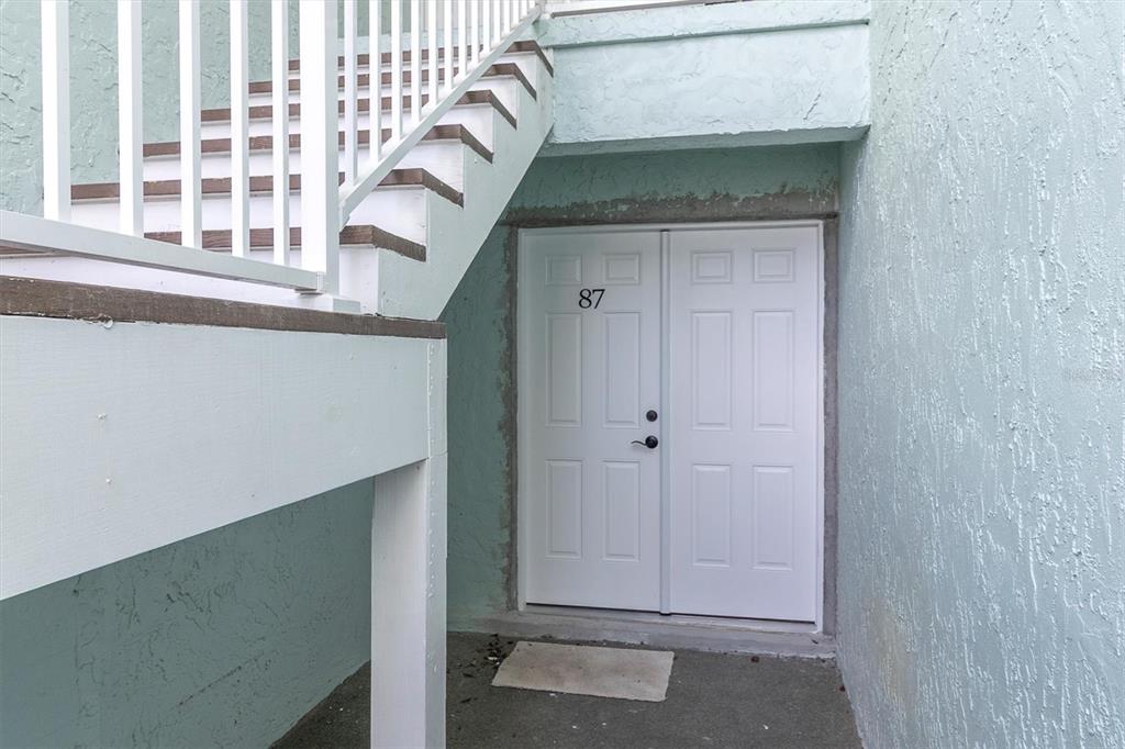 A white double door marked with the number 87 sits beneath an outdoor staircase — 1 WINDRUSH BOULEVARD UNIT 87, INDIAN ROCKS BEACH, FL