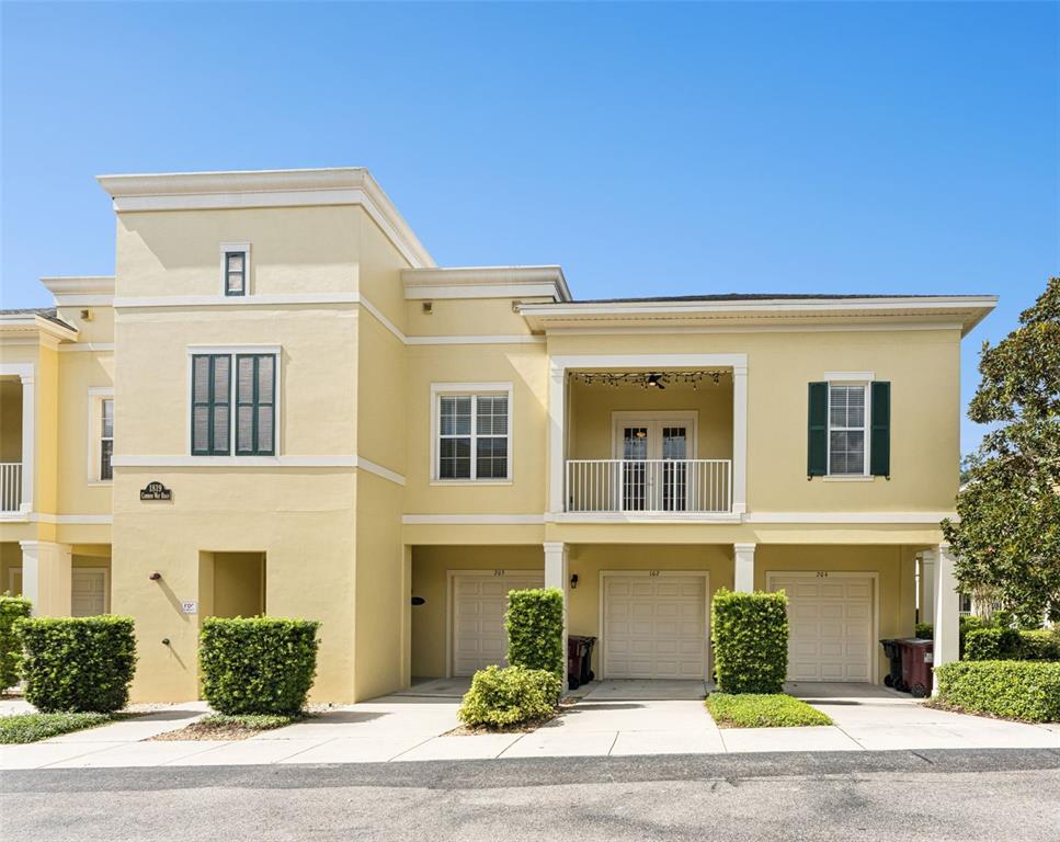 A yellow two-story townhouse features a balcony, three garage doors, and manicured green hedges — 1819 COMMON WAY ROAD UNIT 203, ORLANDO, FL