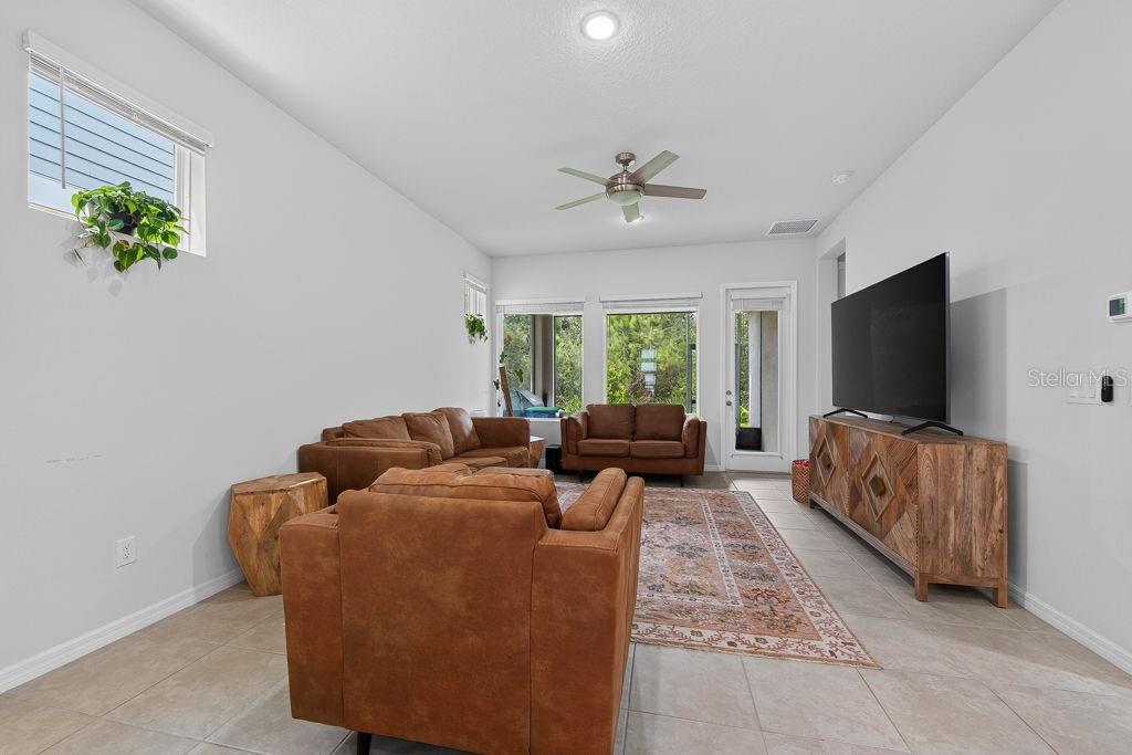 A living room features light tile flooring, brown leather furniture, a large television, and windows — 28583 COZY CREEK DRIVE, WESLEY CHAPEL, FL