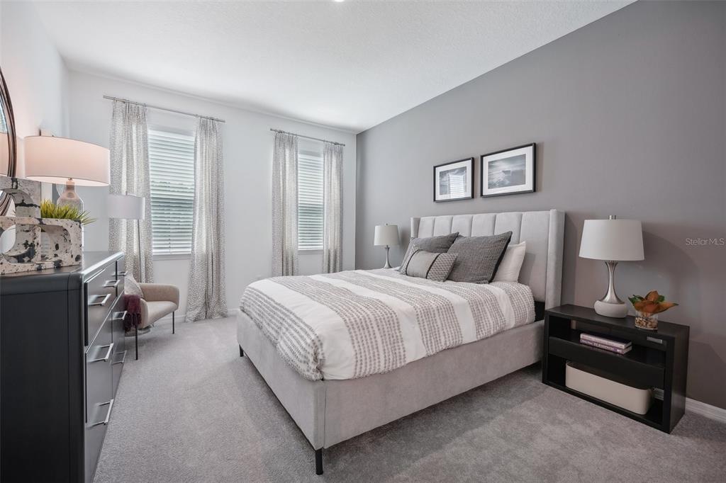 A bedroom features a gray upholstered bed, two windows with curtains, and a black nightstand — 9241 HARRIS HAWK DRIVE, LEESBURG, FL