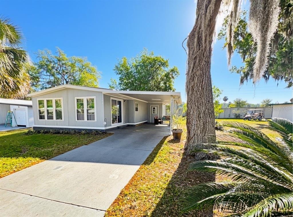 A light gray manufactured home features a concrete driveway, a carport, and large windows — 108 N TIMBER TRAIL, WILDWOOD, FL