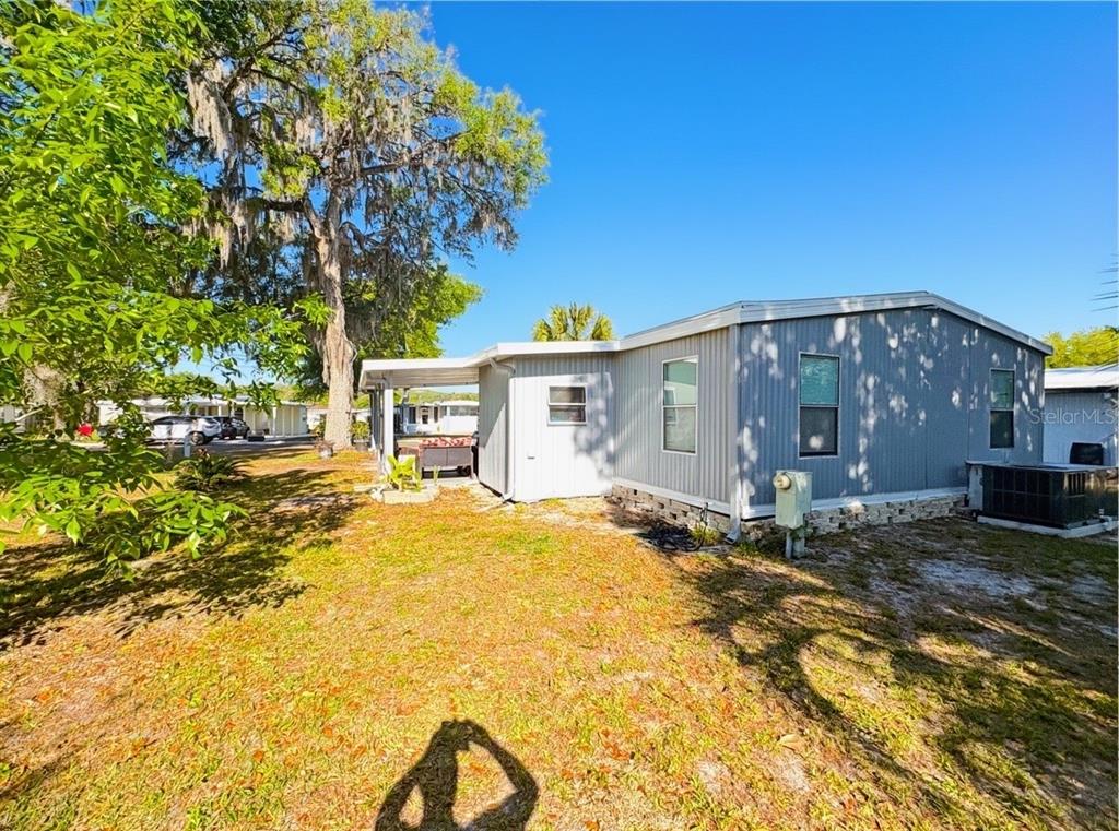 The exterior of a light gray manufactured home features a side entrance and a carport — 108 N TIMBER TRAIL, WILDWOOD, FL