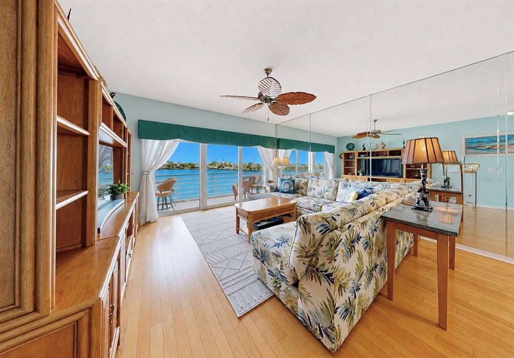 A living room features a floral sectional sofa, wood flooring, mirrored walls, and waterfront views — 4629 MIRABELLA COURT, ST PETE BEACH, FL