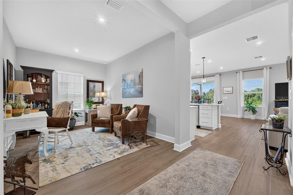 A bright living area with wood-look flooring, two leather armchairs, and an open kitchen beyond — 14076 CRIMSON AVENUE, LAKEWOOD RANCH, FL