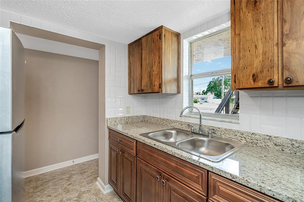 A kitchen with wood cabinets, granite countertops, and white subway tile backsplash leads to a dining area — 5521 BAY BOULEVARD UNIT 104, PORT RICHEY, FL