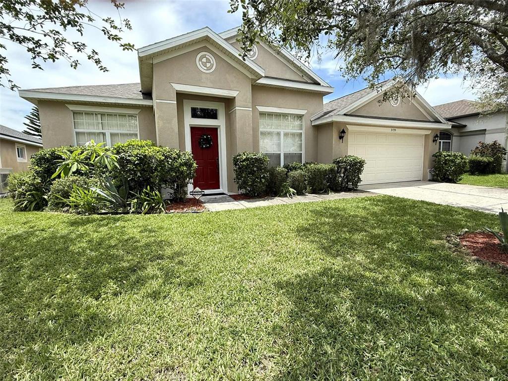 A single-story tan house with a red front door, two-car garage, and landscaped front yard — 3539 MAPLE RIDGE LOOP, KISSIMMEE, FL