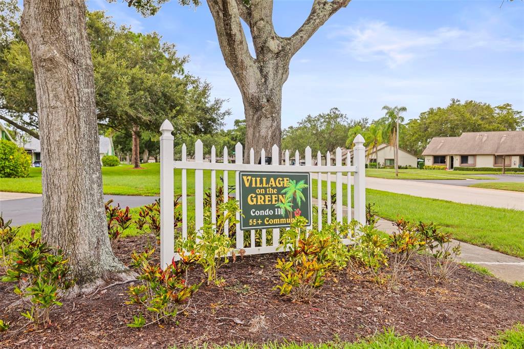 A white picket fence surrounds a sign for Village on the Green 55+ community — 2556 ROYAL PINES CIRCLE UNIT 13-C, CLEARWATER, FL