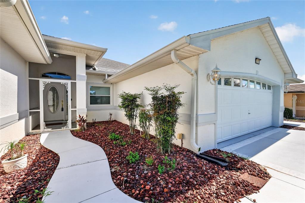 A white house exterior features a curved walkway, red rock landscaping, and a two-car garage — 5049 ST ANDREWS ARCADE, LEESBURG, FL