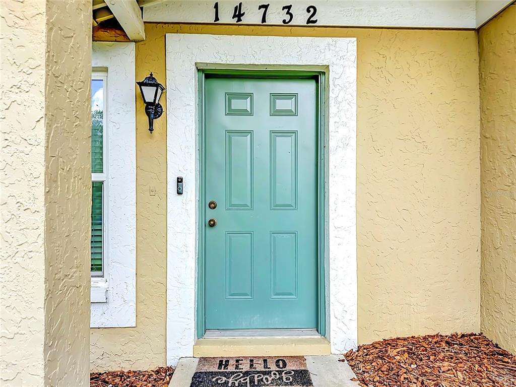 A teal front door with a welcome mat is centered on a tan stucco exterior wall — 14732 PINE GLEN CIRCLE, LUTZ, FL
