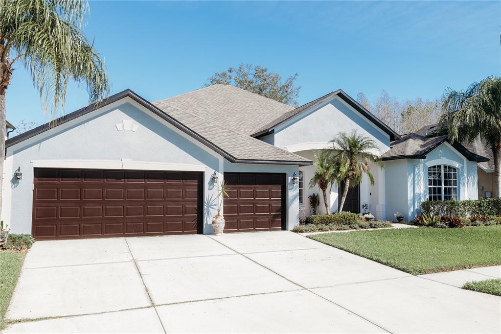 A single-story house with a three-car garage, light blue stucco exterior, and palm trees — 26620 SHOREGRASS DRIVE, WESLEY CHAPEL, FL