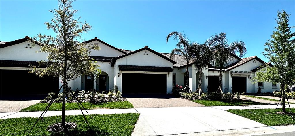 A row of white stucco townhomes features dark garage doors and palm trees under a sky — 5309 KELPER HOOK COURT, WIMAUMA, FL