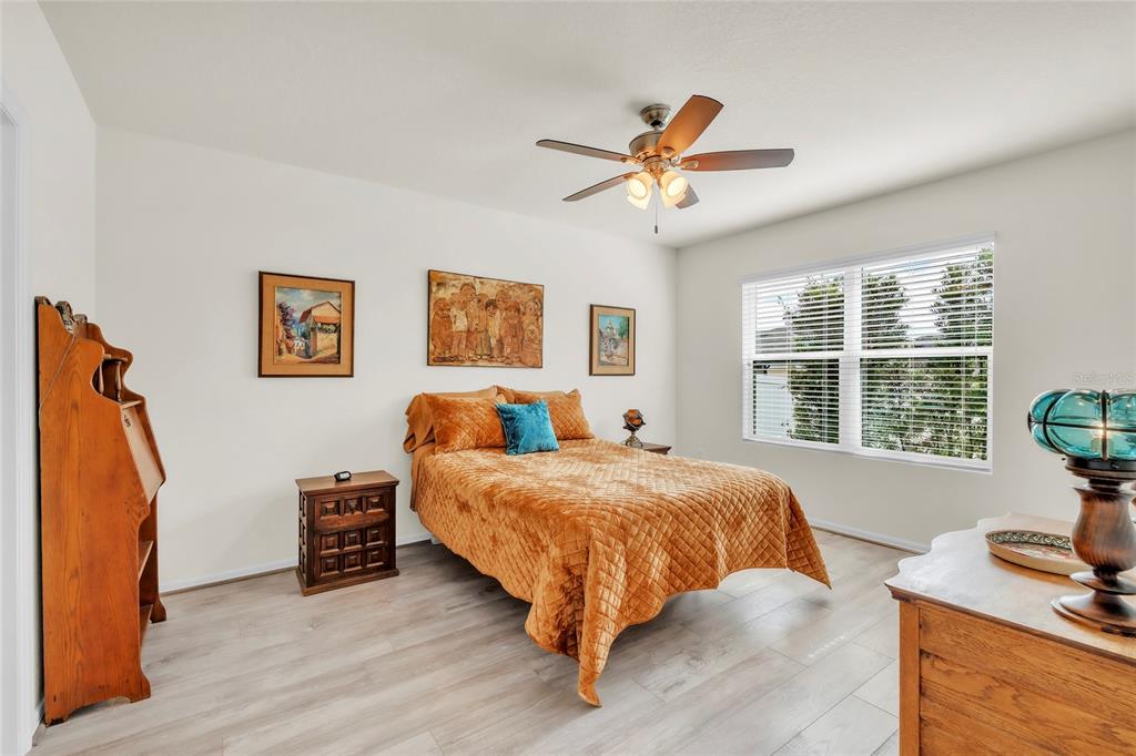 A bedroom features light wood-look flooring, a queen bed with an orange quilt, and a ceiling fan — 9527 SUNSTONE BOULEVARD, THONOTOSASSA, FL
