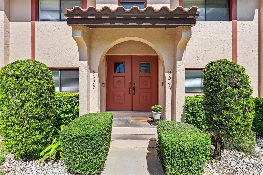 A beige building exterior features a covered entryway with double red doors and manicured hedges — 9543 86TH AVENUE UNIT 9543, SEMINOLE, FL